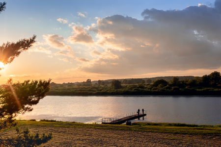 A small river with a wooden Pier is illuminated by the setting sun. The sky is dark and stormy.の写真素材