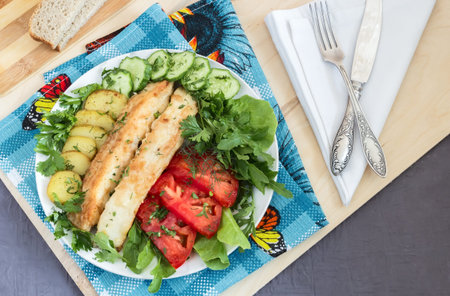 On the table on a plate two slices of fried fish, fried potatoes, cucumbers, tomatoes , lettuce,...