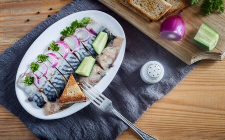 On a wooden table on a napkin plate with slices of salted mackerel, onions, cucumbers, parsley and a piece of bread. Top view, copy spaceの写真素材