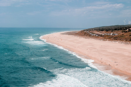 Aerial view of an Atlantic ocean beach with wind turbines in the background in Nazare, Portugal.の写真素材