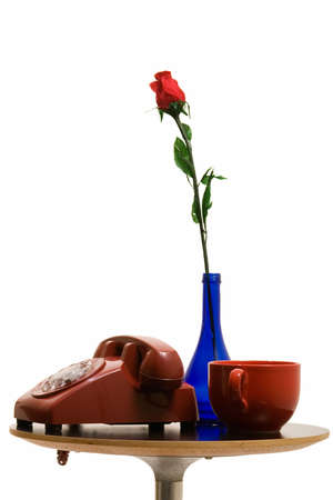 old retro rotary telephone and blue vase with a red rose and a red coffee cup on a small end table over white backgroundの写真素材