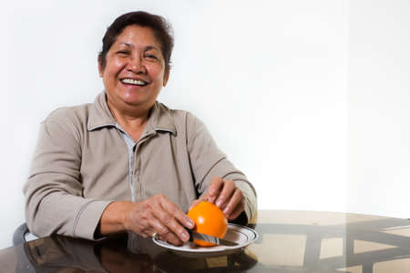 Portrait of a senior asian woman smiling sitting at the kitchen table about to peel and orangeの写真素材
