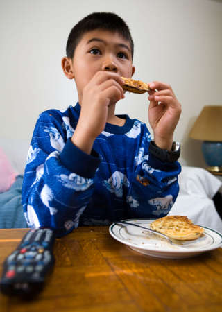Young asian boy eating waffles for breakfast while looking engrossed in the televisionの写真素材