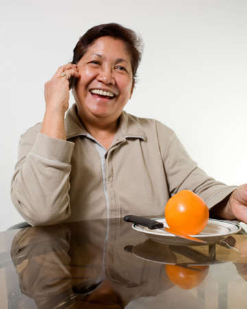 Portrait of a senior asian woman smiling sitting at the kitchen table about to peel and orangeの写真素材