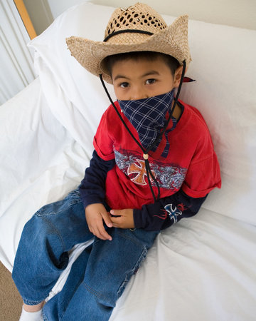 Young asian boy sitting on a white sofa wearing a cowboy hat and a bandana looking at the cameraの写真素材
