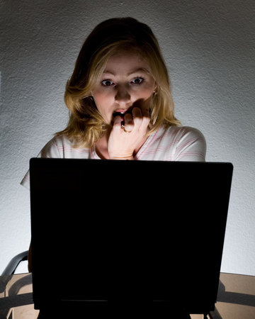 Frightened woman looking at the computer screen in the dark with the light coming from the computer
