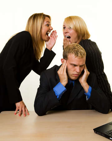 One business man with hands covering hears sitting at desk with two woman coworkers talking behind his backの写真素材