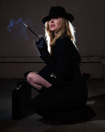 Beautiful blond woman wearing a black trenchcoat and black fedora style hat in a dark alley smoking a cigaretteの写真素材