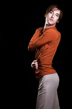 Handsome young brunette Homosexual man wearing a orange turtle neck sweater and beige pants showing curvy figure standing on blackの写真素材