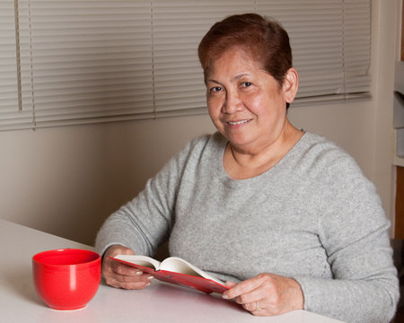 Senior asian woman sitting on a chair relaxing at home smiling holding a red coffee mug and reading a bookの写真素材