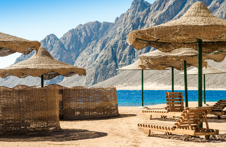 beach umbrellas and wooden lounge chairs on the sand of the beach against the backdrop of the sea and high rocky mountainsの写真素材
