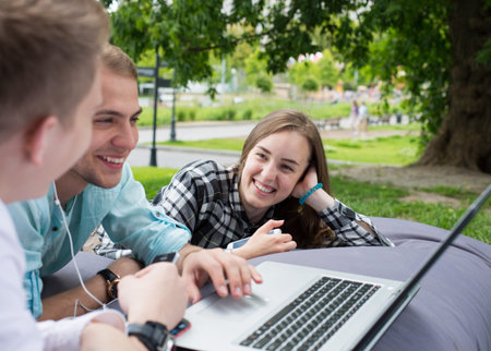 Happy smiling young girl with her male friends chilling on a big cushion outdoors in the park. Friendship or education concept.の写真素材