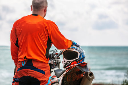 Motorcyclist in a protective suit stands next to motorbike in front of the seaの写真素材