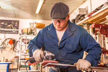 A young man welder in a blue T-shirt, goggles and construction gloves processes metal an angle...
