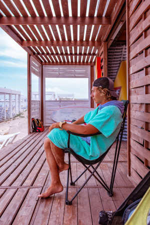 Young attractive guy surfer wearing a bathrobe is sitting on the terrace by the sea, relaxing and...