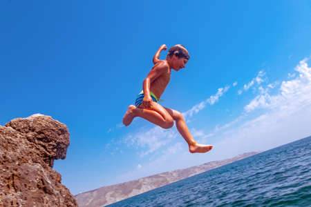 A boy is jumping from the cliff into the sea on a hot summer day. Holidays on the beach. The concept of active tourism and recreationの写真素材