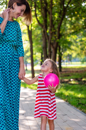 Close-up shoot of pretty small blond long-haired child girl in colorful dress holding trustingly hand of young mother, and rose balloon in the other hand outdoors on blurred colorful sunny summer background.の写真素材