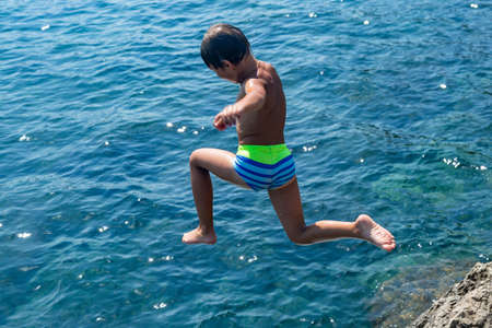 A boy is jumping from the cliff into the sea on a hot summer day. Holidays on the beach. The concept of active tourism and recreationの写真素材