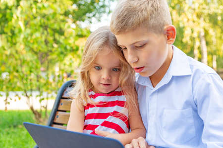 Smiling brother and sister sitting on bench in park and playing on laptopの写真素材
