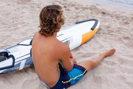Handsome man sits on the beach with white blank surfing board wait for wave to surf spot at sea ocean shore. Concept of sport, fitness, freedom, happiness, new modern life, hipster.の写真素材