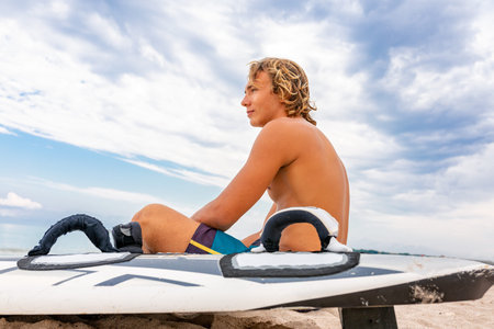 Handsome man sits on the beach with white blank surfing board wait for wave to surf spot at sea...
