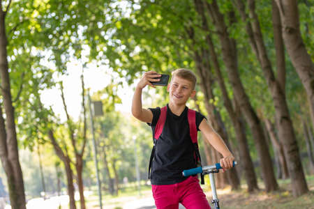 young happy preteen boy riding a scooter in warm sunny summer day in the park and taking selfie on smartpone while riding on scooterの写真素材
