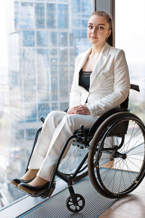 Confident happy businesswoman in wheelchair against the background of a panoramic window overlooking the skyscrapers and a big city., she is smiling at camera, disability overcoming conceptの写真素材