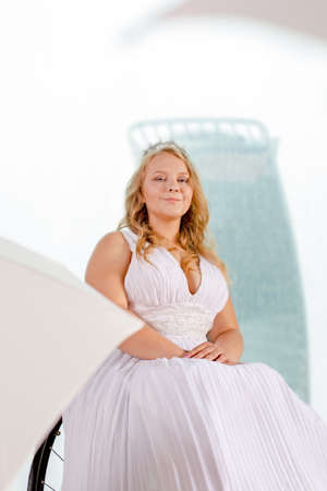 Beautiful disabled bride posing in a wheelchair against the background of a panoramic window overlooking the skyscrapers and a big city, she is smiling at camera, disability overcoming conceptの写真素材