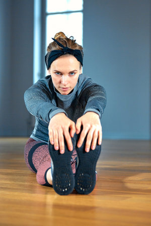Young beautiful woman in sportswear doing stretching while sitting on the floor in front of window...