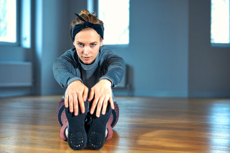 Young beautiful woman in sportswear doing stretching while sitting on the floor in front of window...