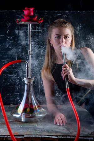 Young, beautiful woman smokes a hookah with red roses. It produces smoke from his mouth. The pleasure of smoking. rose aroma hookahの写真素材