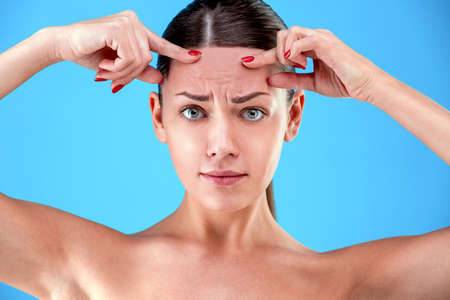 Signs of aging. Portrait of shocked young woman is looking at camera with disappointedly while touching her wrinkles on forehead on blue backgroundの写真素材