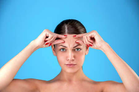 Signs of aging. Portrait of shocked young woman is looking at camera with disappointedly while touching her wrinkles on forehead on blue backgroundの写真素材