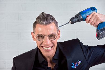 Portrait of crazy funny young man in black suit smiling at camera with a drill holds at his temple....