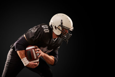 American football player in dark uniform with the ball is preparing to attack on a black background.の写真素材