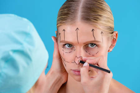 Beautician draw correction lines on woman face. Before plastic surgery operetion. Isolated on blueの写真素材