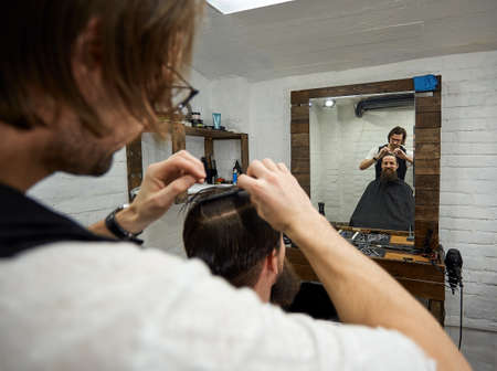 Brutal guy in modern Barber Shop. Hairdresser makes hairstyle a man with a long beard. Master hairdresser does hairstyle by scissors and combの写真素材