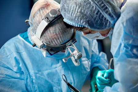 Surgeon and his assistant performing cosmetic surgery in hospital operating room. Surgeon in mask wearing loupes during medical procadure.の写真素材