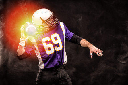 American football player holding ball in his hands in smoke. Black background, copy space. The...