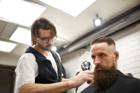Brutal guy in modern Barber Shop. Hairdresser makes hairstyle a man with a long beard. Master hairdresser does hairstyle by scissors and combの写真素材
