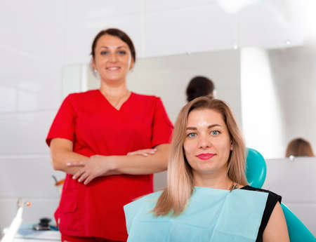 Dentist finishing successful teeth restoration, black haired doctor, in green mask, giving to happy patient mirror, beautiful long-haired woman looking at pristine white teeth with great pleasureの写真素材