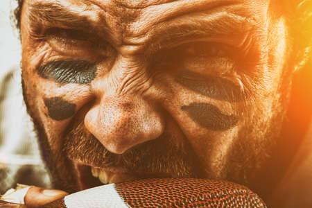 Closeup portrait of an aggressive American football player. Aggressive player bites his ball. The concept of American football, sports anger, close-up, special illumination, the effect of film grain.の写真素材