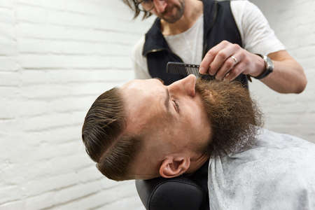 Brutal guy in modern Barber Shop. Hairdresser makes hairstyle a man with a long beard. Master hairdresser does hairstyle with hair clipperの写真素材