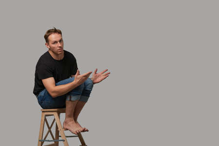 A man in a black T-shirt and jeans sitting on a chair shows with his hands on your text or your advertisement on a gray background. Concept banner for advertising, copy space, gray background, commercial design.の写真素材