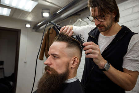 Master cuts hair and beard of men in the barbershop, hairdresser makes hairstyle for a young manの写真素材