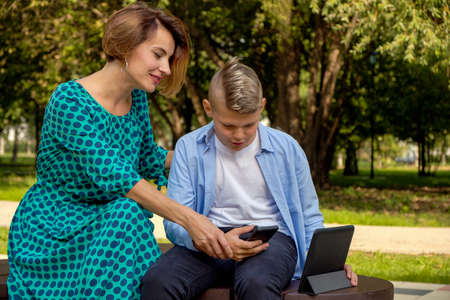 Mom and son using tablet and smartphone while sitting on nature. The family plays computer or searches the Internet, electronic devices and children in modern life.の写真素材
