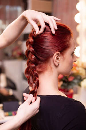 Beautiful, red-haired girl with long hair, hairdresser weaves a French braid, in a beauty salon. Professional hair care and creating hairstyles.の写真素材
