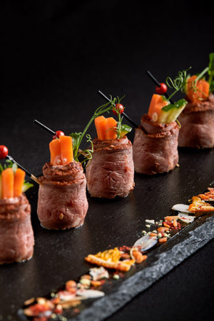 Mini roast beef rolls with vegetables, on a black slate plate, on a black background. Fusion food...