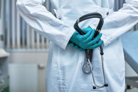 Doctor holding stethoscope behind her back, waist-high portrait on clinic background.の写真素材