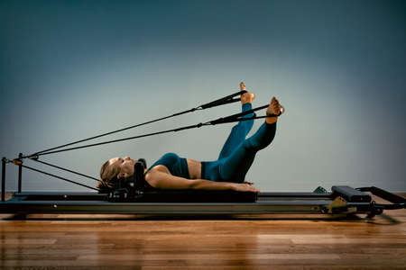 Young girl doing pilates exercises with a reformer bed. Beautiful slim fitness trainer on reformer gray background, low key, art light. Fitness concept.の写真素材